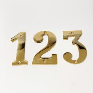 Puede incluir: Tres números de casa de metal de color dorado, 1, 2 y 3, están dispuestos en fila sobre un fondo blanco.