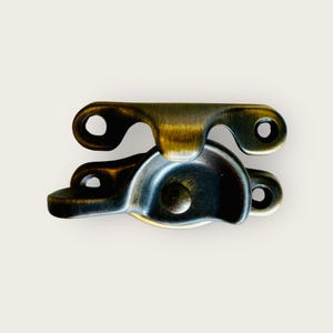 Może przedstawiać: Zamek do okna w kolorze mosiądzu o zaokrąglonym, okrągłym kształcie. Zamek ma dwa ramiona połączone zawiasem.