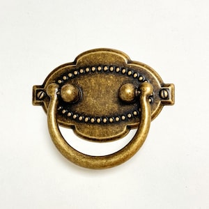 Op de afbeelding: Een messingkleurige metalen kastgreep met een decoratief ovaal ontwerp en een ringvormige handgreep. De greep heeft een verweerde afwerking en een parelrand.