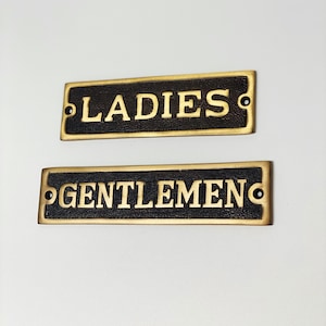 May include: Two brass signs with black backgrounds and gold lettering. The top sign reads "LADIES" and the bottom sign reads "GENTLEMEN".