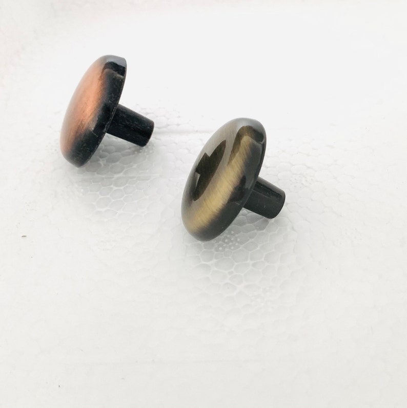 May include: Two round, decorative cabinet knobs with a black base and a metallic finish. One knob is copper colored and the other is gold colored.