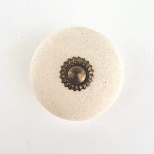 May include: A round, off-white ceramic cabinet knob with a textured surface. The knob features a decorative, antique brass-colored metal center with a sunburst design. Ideal for adding a vintage touch to furniture.