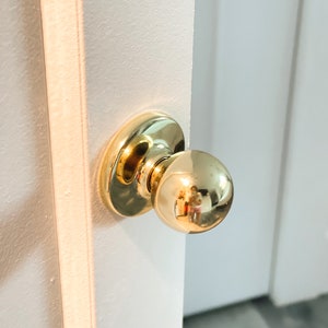 Puede incluir: Un pomo de puerta de oro pulido con un mango redondo. El pomo está montado en una puerta blanca.