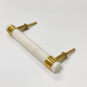 May include: A white marble cabinet pull with gold-colored accents. The cylindrical handle is flanked by gold bands and attached to the cabinet with gold-colored screws. The hardware is designed for furniture and home decor.