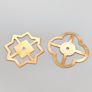 Puede incluir: Dos piezas decorativas de metal dorado con intrincados diseños recortados. Una pieza es una estrella con un cuadrado en el centro, la otra es una forma de trébol de cuatro hojas.