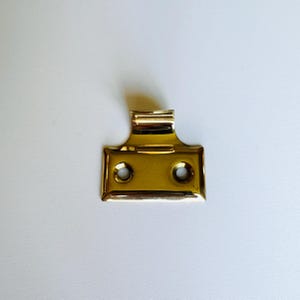 May include: A brass window sash lift with two holes. The lift has a curved top and a rectangular base.