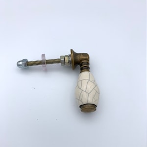 Puede incluir: Un pomo de armario de cerámica blanca con acabado craquelado y base de latón. El pomo tiene un tornillo y una arandela.