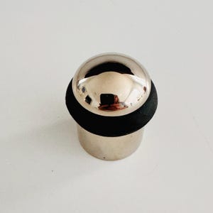 May include: A chrome door stop with a spherical top and a cylindrical base. The top is reflective, and the base is surrounded by a black rubber ring. The door stop is designed to prevent doors from slamming shut.