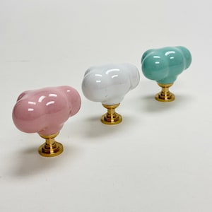 Può includere: Tre pomelli per armadi in ceramica a forma di nuvola. I pomelli sono rosa, bianco e azzurro. Ogni pomello ha una base in metallo dorato.
