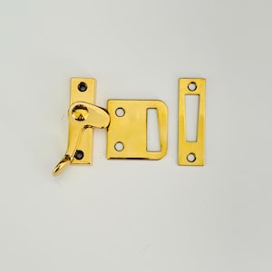 Puede incluir: Pestillo de ventana dorado con una manija de palanca y una placa de golpe rectangular. El pestillo está diseñado para asegurar una ventana desde el interior.