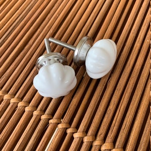 Peut inclure: Deux boutons de meuble en céramique blanche avec un design côtelé en forme de citrouille. Chaque bouton a une base et une vis en métal argenté. Les boutons sont présentés sur une surface en bambou tressé brun.
