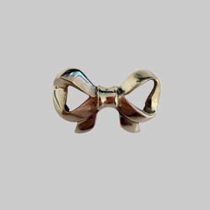 May include: A shiny, silver-colored bow-shaped brooch. The brooch has a detailed design with curved loops and a central knot. The metal reflects light, creating highlights and shadows. The bow is a classic accessory, suitable for various outfits.