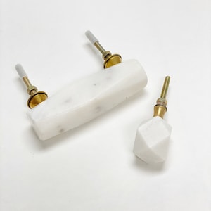 May include: Three white marble cabinet knobs with brass fittings. One knob is rectangular, and the other is a faceted geometric shape. The knobs are designed for easy installation and add a touch of elegance to any furniture piece.