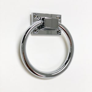 May include: A polished chrome towel ring with a rectangular backplate. The ring is circular and attached to the backplate with a rectangular connector. The hardware is mounted on a white surface.
