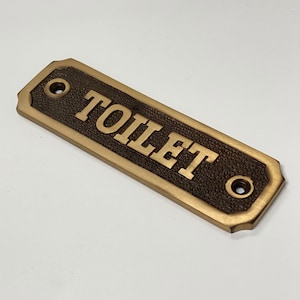 Könnte beinhalten: Ein goldfarbenes Metallschild mit einer strukturierten Oberfläche und dem Wort "TOILET" in erhabenen Buchstaben. Das Schild hat zwei kleine Löcher an beiden Seiten.
