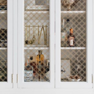 May include: White cabinet with glass doors and gold wire mesh inserts. The cabinet has two shelves and is filled with various bottles and glasses. The cabinet has two doors on the bottom that are closed.