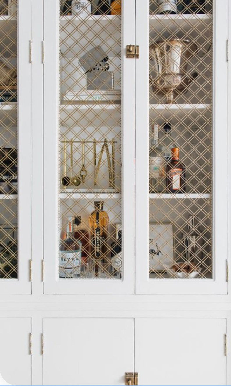 May include: White cabinet with glass doors and gold wire mesh inserts. The cabinet has two shelves and is filled with various bottles and glasses. The cabinet has two doors on the bottom that are closed.