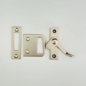 May include: A silver-toned window latch with a lever handle and a strike plate. The latch has two screws and a small hole for a key.