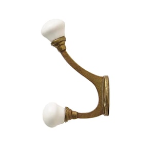 Könnte beinhalten: Ein dekorativer Wandhaken im Vintage-Stil. Der Haken besteht aus messingfarbenem Metall und hat zwei weiße Keramikknöpfe. Der Haken ist gebogen und hat eine runde Basis zur Montage.