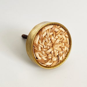 May include: A round, brass cabinet knob with a woven, natural fiber center. The knob has a screw on the back for attaching to a cabinet door.