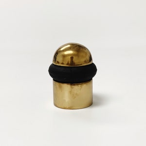 Puede incluir: Un tope de puerta dorado con una base de goma negra. El tope de puerta tiene forma de esfera.