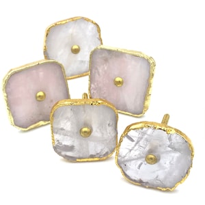 Peut inclure: Ensemble de quatre boutons de cabinet carrés en cristal rose et blanc avec une bordure dorée. Chaque bouton a un petit bouton doré au centre.