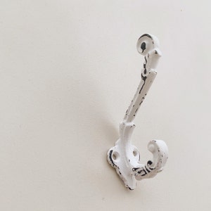 Könnte beinhalten: Weißer Metallhaken mit einem dekorativen, gerollten Design. Der Haken ist an einer weißen Wand montiert.