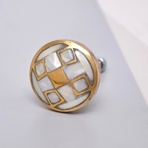 Puede incluir: Un pomo redondo de latón para armario con un diseño geométrico hecho de incrustaciones de nácar blanco.