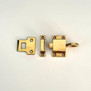 Puede incluir: Un pestillo de metal dorado con un asa de bucle. El pestillo tiene tres partes: una placa de golpe, un retenedor y un pestillo con asa.