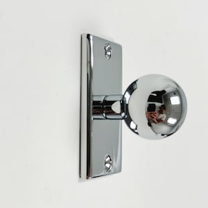 May include: A shiny chrome door knob with a spherical handle. It's fixed to a rectangular backplate with screw holes. The spherical handle reflects the surroundings. The door hardware is designed for interior doors.
