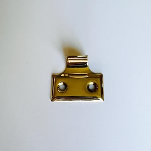May include: A brass window sash lift with two holes. The lift has a curved top and a rectangular base.