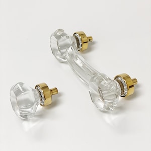May include: A pair of clear glass cabinet knobs with gold metal accents. The knobs are faceted and have a curved shape.
