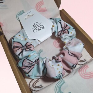 May include: A patterned scrunchie in a gift box. The scrunchie has a light blue background with pink bows and gold dots. The gift box contains tissue paper with a rainbow design and a tag that reads "Lil Boo Crafts."