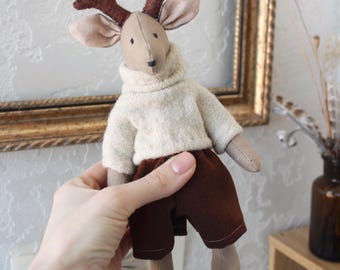 May include: A handmade plush deer doll with brown antlers, tan ears, and a beige sweater. The deer wears brown corduroy pants and has tan feet. The doll is being held in front of a decorative gold framed picture.