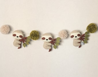 May include: A garland with three brown sloth plushies holding onto green branches. The garland is strung with beige and green pom-poms.