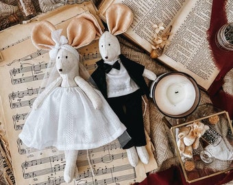 May include: Two handmade fabric mouse dolls dressed in wedding attire. The bride mouse wears a white dress with lace trim and a veil. The groom mouse wears a black suit with a white bow tie. The dolls are sitting on a sheet of music with a vintage book, a bowl of buttons, and a candle in the background.