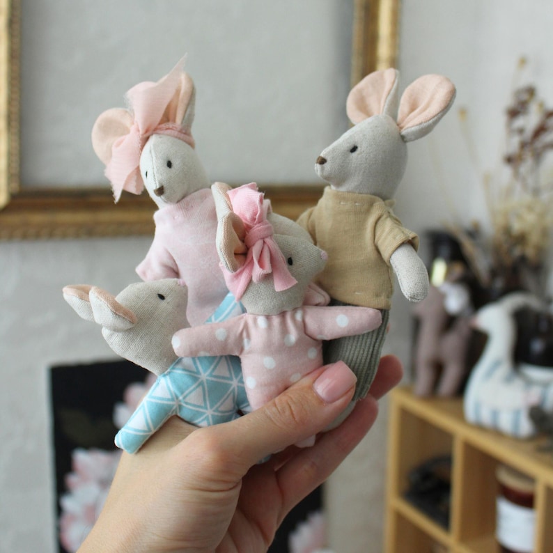 May include: A collection of handmade fabric mouse dolls. The dolls are dressed in various outfits, including pink and blue patterned clothing, and adorned with bows. The mice have long ears and are held in a hand.