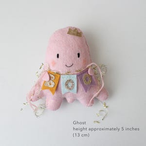 May include: A pink felt ghost with a gold crown and a banner that reads "BOO". The ghost is approximately 5 inches (13 cm) tall. The banner has gold, blue, and purple pennants with glitter accents. The ghost has a smiling face.