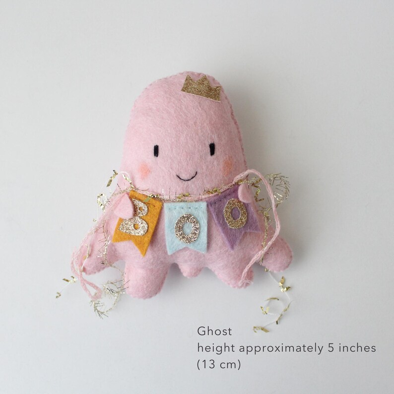 May include: A pink felt ghost with a gold crown and a banner that reads "BOO". The ghost is approximately 5 inches (13 cm) tall. The banner has gold, blue, and purple pennants with glitter accents. The ghost has a smiling face.