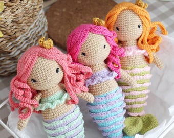 May include: Three handmade crochet mermaid dolls with pink, orange, and pink hair, each wearing a gold crown. The dolls have colorful striped tails in shades of blue, green, and purple. They are displayed in a white wire basket.
