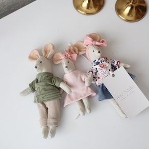 May include: Three handmade fabric mouse dolls. One mouse wears a green shirt and tan pants, another a pink dress, and the third a floral top and blue skirt. Each mouse has large ears and a pink bow.