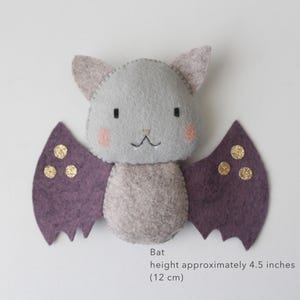 May include: A handmade felt bat toy in shades of gray and purple. The bat has a light gray body, with purple wings adorned with gold glitter accents. The face features small black eyes, rosy cheeks, and a stitched smile. Height approximately 4.5 inches.