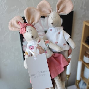 May include: Two plush mouse dolls with large, light pink ears. One mouse wears a pink bow, floral print top, and pink skirt. The other mouse wears a white shirt with mushroom graphics. Both dolls have a tag that reads "tiny & tale."