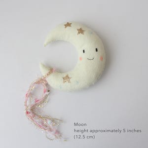May include: A plush, off-white crescent moon with a smiling face and rosy cheeks. The moon is decorated with gold glitter stars and small blue stars. Pink and gold fringe hangs from the moon's tip. The moon is approximately 5 inches (12.5 cm) tall.