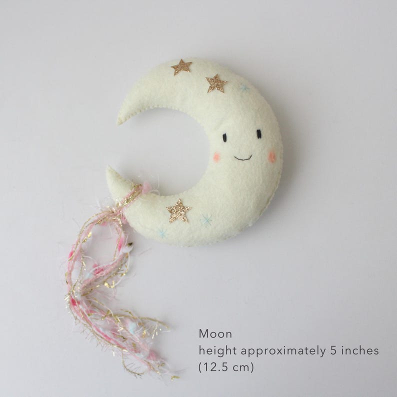 May include: A plush, off-white crescent moon with a smiling face and rosy cheeks. The moon is decorated with gold glitter stars and small blue stars. Pink and gold fringe hangs from the moon's tip. The moon is approximately 5 inches (12.5 cm) tall.