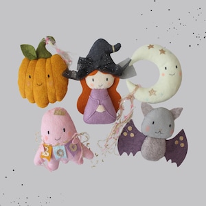May include: A collection of handmade Halloween-themed felt ornaments. Includes a smiling orange pumpkin, a witch with a black hat and purple dress, a pink ghost, a crescent moon with stars, and a gray bat with purple wings.
