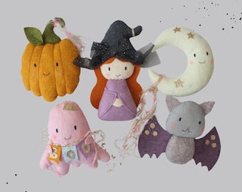 May include: A collection of handmade Halloween-themed felt ornaments. Includes a smiling orange pumpkin, a witch with a black hat and purple dress, a pink ghost, a crescent moon with stars, and a gray bat with purple wings.