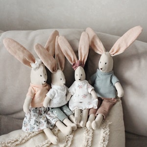 May include: Four fabric bunny dolls with long ears. Each bunny is dressed in a different outfit, including a floral dress, a pink top and skirt, a white top and trousers, and a blue top and trousers. They are sitting on a cream-coloured cushion.