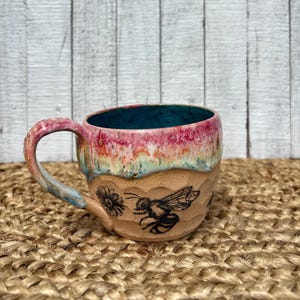May include: A handmade ceramic mug with a bee and flower design. The mug features a textured, tan-colored base with a black illustration of a bee and a flower. The rim is a gradient of pink, yellow, and blue, with a teal interior. The handle is pink and blue.