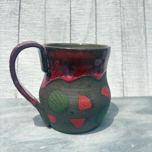 Peut inclure: Mug en céramique fait à la main avec une base gris foncé et un bord rouge vif. Le mug présente des cercles verts et des formes géométriques rouges. La anse et le bord ont une finition brillante.